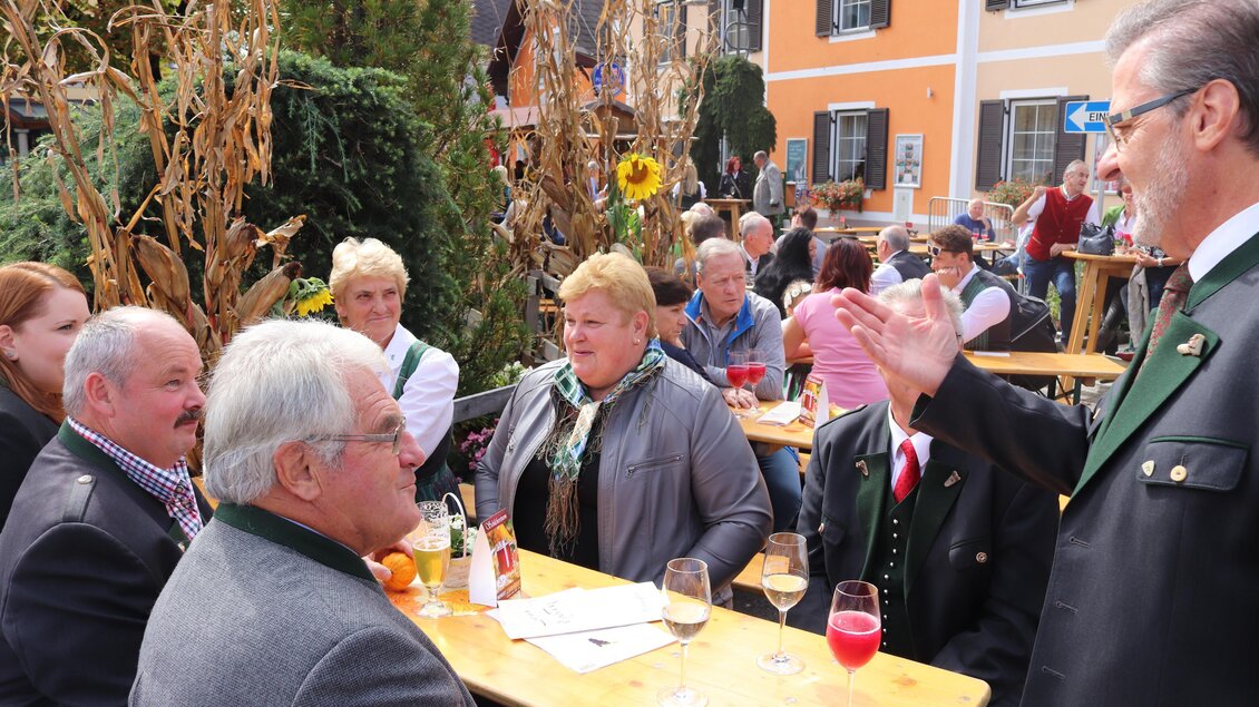 Eine Gruppe von Menschen sitzt an Tischen im Freien und unterhält sich. Es sind Festlichkeiten in einem ländlichen Ambiente mit herbstlicher Dekoration zu sehen. | © Gem. Mooskirchen-Robert Cescutti