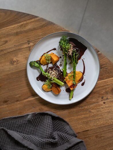 A plate with sautéed vegetables, including broccoli and pumpkin, in a delicious sauce. Next to the plate, there is a dish towel on a wooden table. | © Das Zeitwert