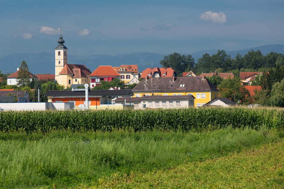 Eine malerische Landschaft mit einem Dorf im Hintergrund. Die Kirche und die bunten Häuser sind gut sichtbar, umgeben von grünen Feldern. | © Region Graz-Harry Schiffer