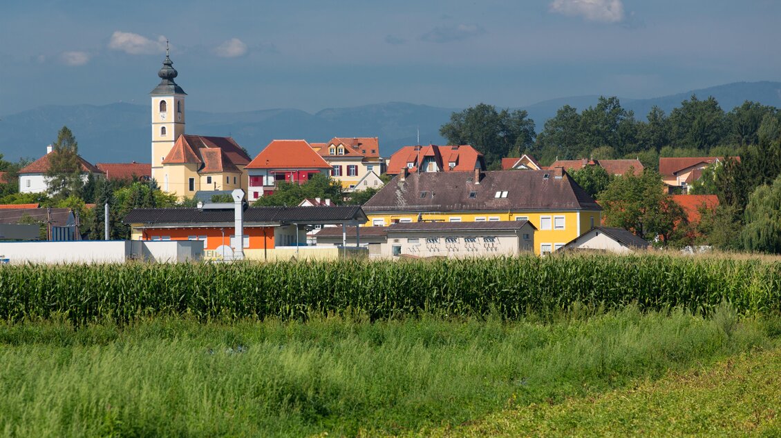 Eine malerische Landschaft mit einem Dorf im Hintergrund. Die Kirche und die bunten Häuser sind gut sichtbar, umgeben von grünen Feldern. | © Region Graz-Harry Schiffer