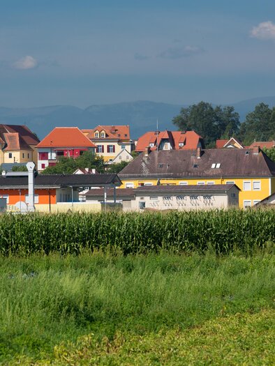 Eine malerische Landschaft mit einem Dorf im Hintergrund. Die Kirche und die bunten Häuser sind gut sichtbar, umgeben von grünen Feldern. | © Region Graz-Harry Schiffer