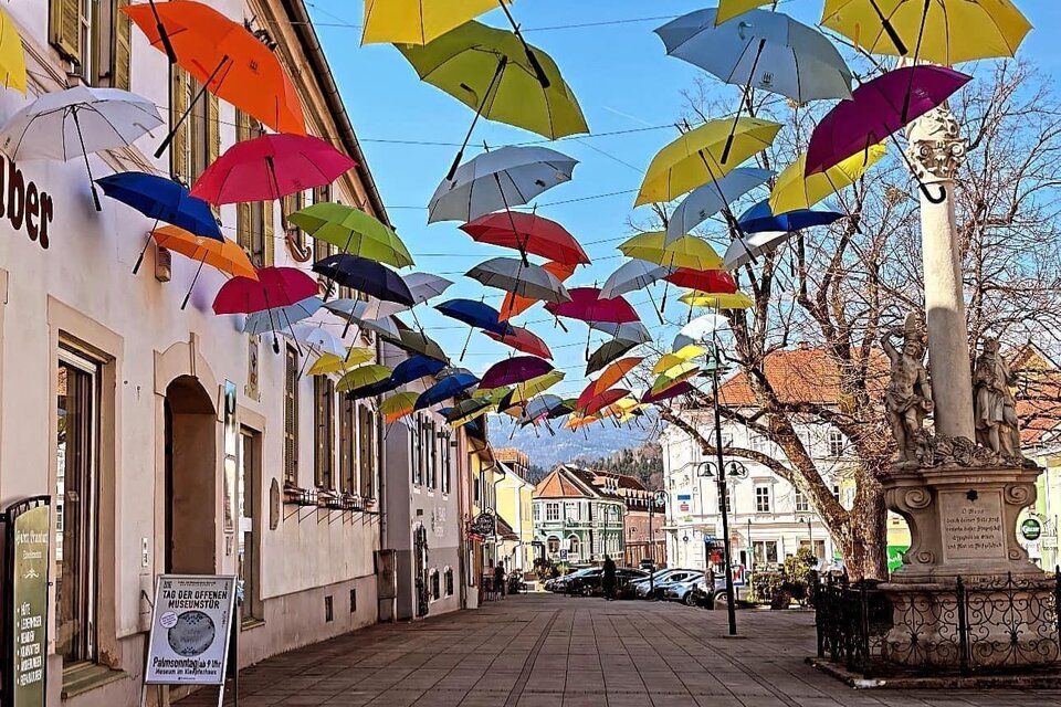 Eine bunte Straße mit vielen Regenschirmen, die über den Gehweg hängen. Die Gebäude sind hell und die Stimmung ist fröhlich. | © TVB Südsteiermark