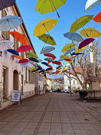 A colorful street with many umbrellas hanging over the sidewalk. The buildings are bright and the atmosphere is cheerful. | © TVB Südsteiermark