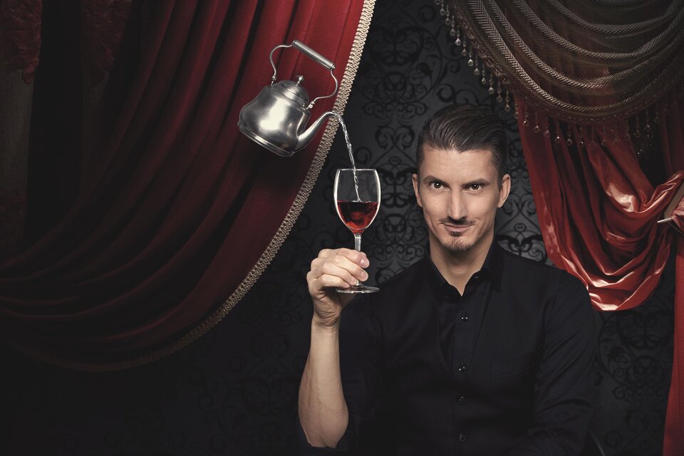 Ein Mann hält ein Glas Wein in einer eleganten Umgebung. Im Hintergrund sind rote Vorhänge und eine Teekanne zu sehen. | © Felicitas Matern - Feelimage