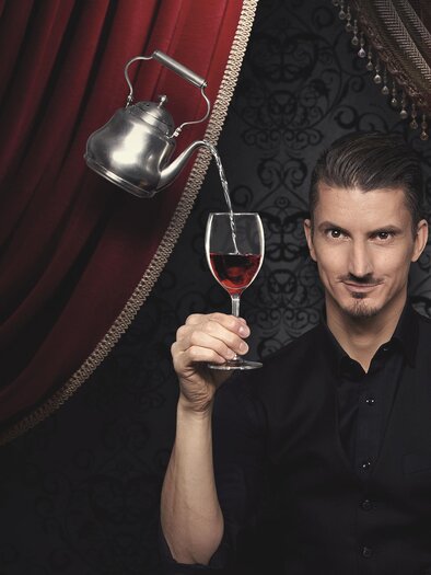 Ein Mann hält ein Glas Wein in einer eleganten Umgebung. Im Hintergrund sind rote Vorhänge und eine Teekanne zu sehen. | © Felicitas Matern - Feelimage