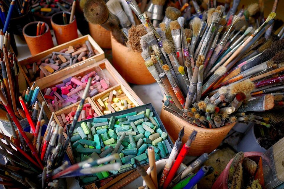 Ein Tisch voller Künstlerbedarf mit Pinsel, Stiften und pastellfarbenen Kreiden. Die Materialien sind in verschiedenen Behältern organisiert und zeigen eine kreative Arbeitsumgebung. | © pixabay.com