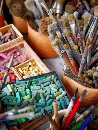 Ein Tisch voller Künstlerbedarf mit Pinsel, Stiften und pastellfarbenen Kreiden. Die Materialien sind in verschiedenen Behältern organisiert und zeigen eine kreative Arbeitsumgebung. | © pixabay.com