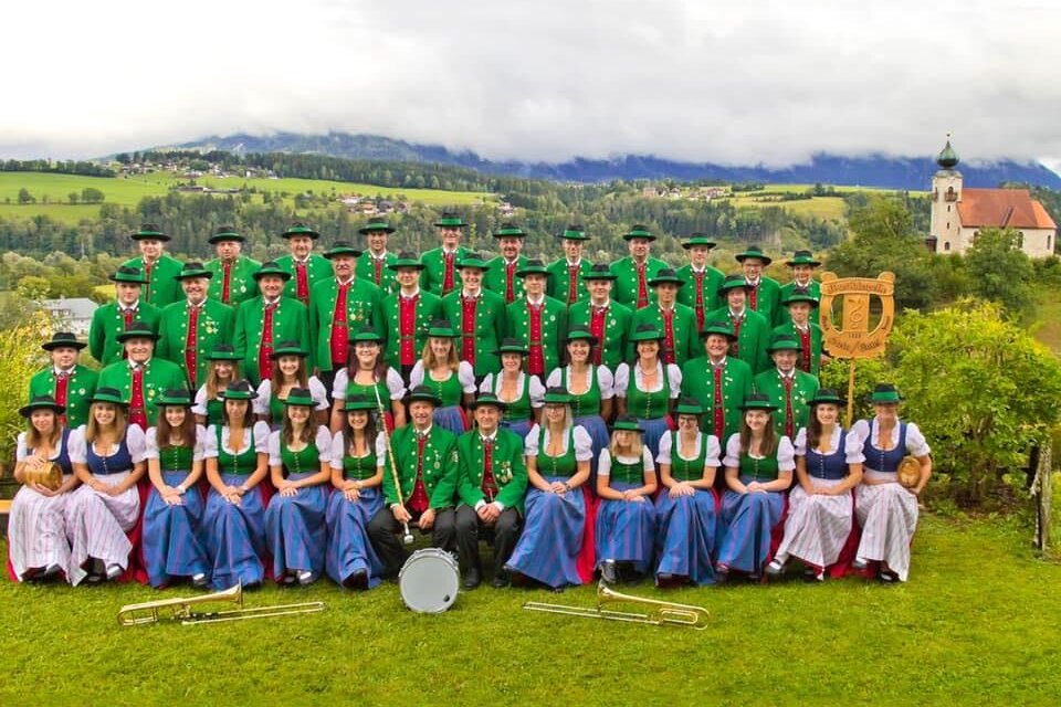 Eine Gruppe von Musikern in traditionellen Trachten steht auf einer Wiese. Im Hintergrund sind Berge und ein historisches Gebäude zu sehen.