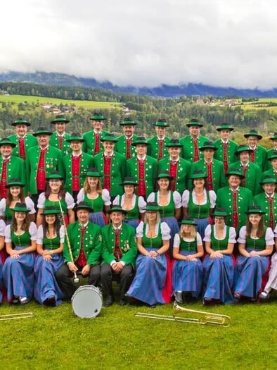 Eine Gruppe von Musikern in traditionellen Trachten steht auf einer Wiese. Im Hintergrund sind Berge und ein historisches Gebäude zu sehen.