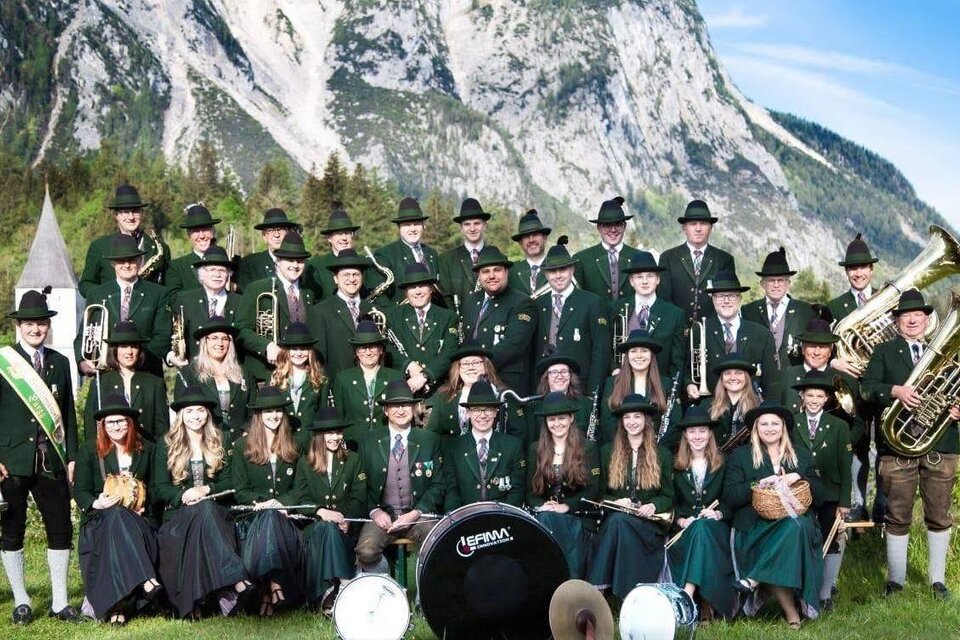 Eine große Gruppe von Musikern in traditionellen Trachten steht vor majestätischen Bergen. Sie halten Musikinstrumente und posieren fröhlich in der Natur. | © tmk pürgg
