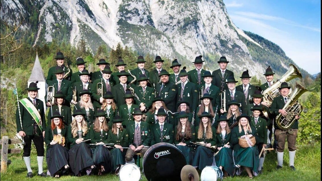 Eine große Gruppe von Musikern in traditionellen Trachten steht vor majestätischen Bergen. Sie halten Musikinstrumente und posieren fröhlich in der Natur. | © tmk pürgg