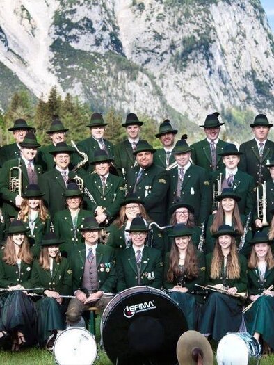 Eine große Gruppe von Musikern in traditionellen Trachten steht vor majestätischen Bergen. Sie halten Musikinstrumente und posieren fröhlich in der Natur. | © tmk pürgg