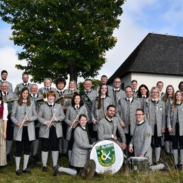 Eine große Gruppe von Menschen steht vor einer Kapelle. Die Mitglieder tragen traditionelle Kleidung und halten Instrumente in den Händen.