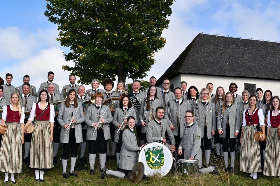 Eine große Gruppe von Menschen steht vor einer Kapelle. Die Mitglieder tragen traditionelle Kleidung und halten Instrumente in den Händen.