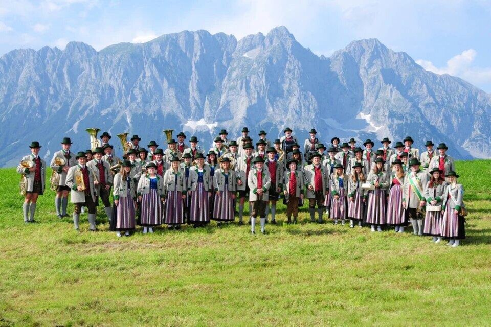 Eine Gruppe von Menschen in traditioneller Tracht steht vor einer majestätischen Berglandschaft. Die Atmosphäre ist festlich und farbenfroh. | © Musikverein Öblarn
