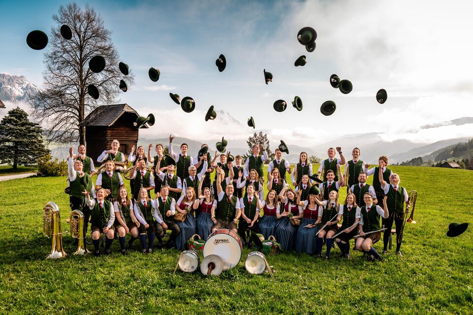 Eine fröhliche Gruppe von Musikern steht auf einer Wiese. Sie tragen traditionelle Trachten und werfen ihre Hüte in die Luft. | © David Stocker
