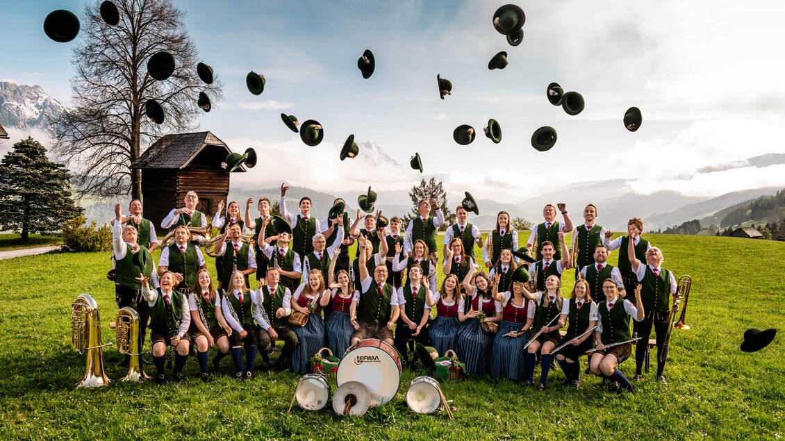 Eine fröhliche Gruppe von Musikern steht auf einer Wiese. Sie tragen traditionelle Trachten und werfen ihre Hüte in die Luft. | © David Stocker