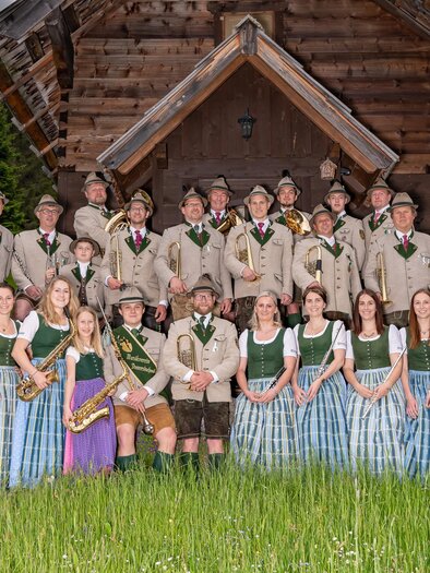 Gruppenfoto eines Musikvereins vor einer großen Holzhütte in einer grünen Berglandschaft. Etwa zwei Dutzend Menschen stehen in mehreren Reihen. Die Männer tragen traditionelle Tracht mit Hüten und halten Blasinstrumente wie Trompeten, Hörner und Tuben. Die Frauen tragen lange Dirndl in Grün, Weiß und Blau. Im Vordergrund wächst hohes Gras, im Hintergrund sind Wald und Berge zu sehen.