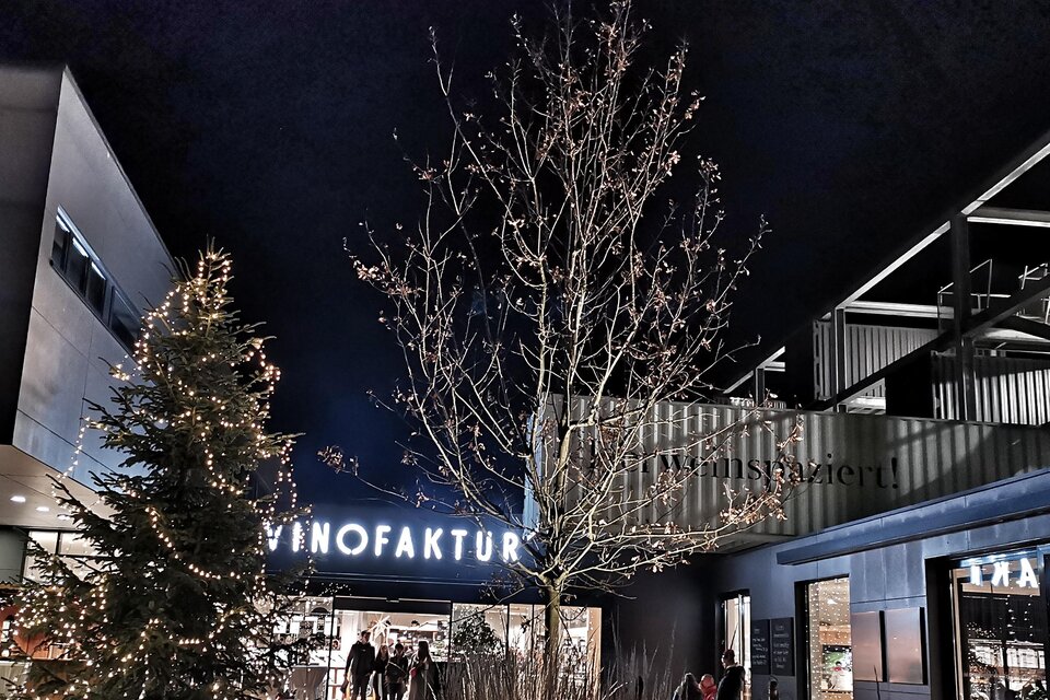 Eine festlich beleuchtete Szenerie mit einem großen Weihnachtsbaum und einem Gebäude mit dem Schriftzug "VINOFAKTUR". Die Atmosphäre ist abends und das Bild strahlt eine einladende Stimmung aus. | © Vinofaktur im Genussregal