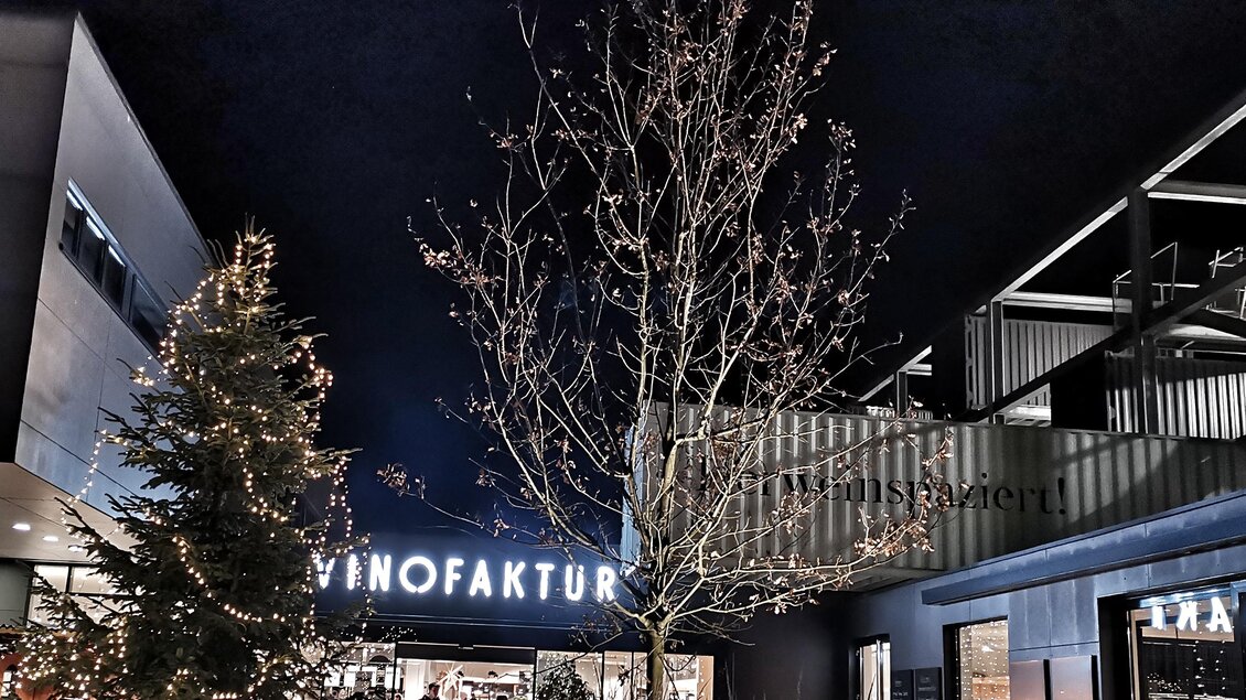 Eine festlich beleuchtete Szenerie mit einem großen Weihnachtsbaum und einem Gebäude mit dem Schriftzug "VINOFAKTUR". Die Atmosphäre ist abends und das Bild strahlt eine einladende Stimmung aus. | © Vinofaktur im Genussregal