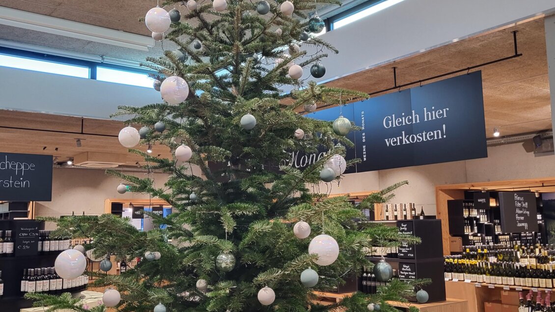 Ein geschmückter Weihnachtsbaum mit weißen und blauen Kugeln steht im Geschäft. Im Hintergrund sind Regale mit Produkten sichtbar. | © Vinofaktur im Genussregal