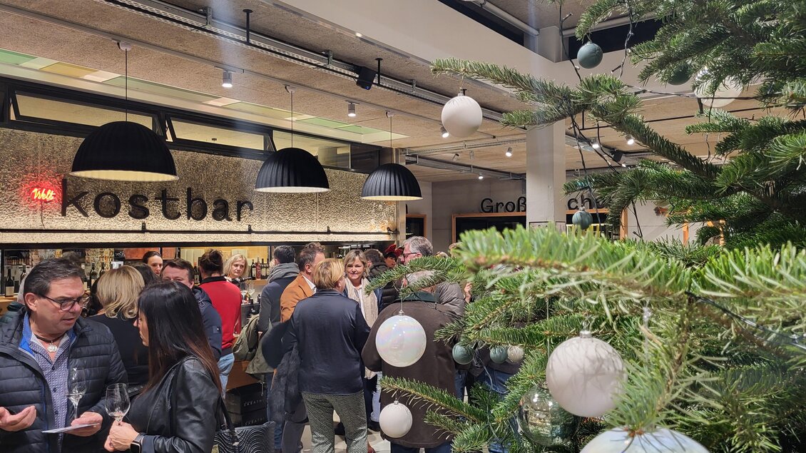 Ein festlich dekorierter Raum mit einer Menschenmenge, die sich am Buffet versammelt. Im Vordergrund steht ein Tannenbaum mit bunten Kugeln. | © Vinofaktur im Genussregal