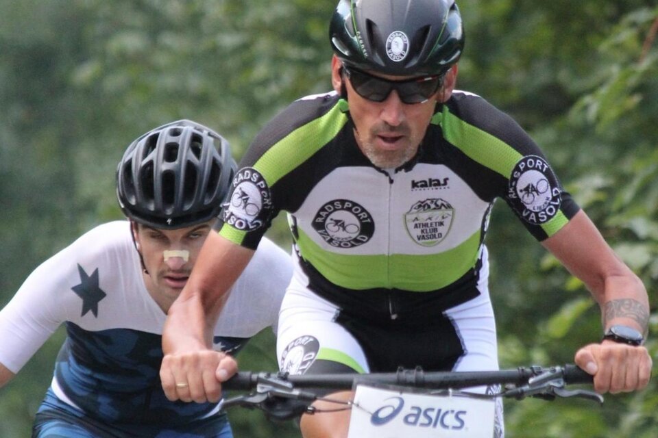 Zwei Radfahrer in sportlicher Bekleidung fahren auf einem Waldweg. Der Fokus liegt auf dem vorneliegenden Fahrer, der konzentriert und schnell unterwegs ist.