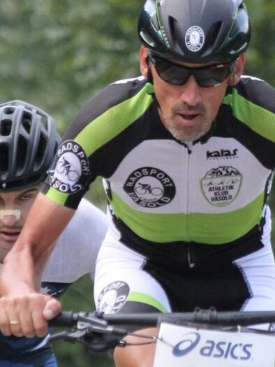 Zwei Radfahrer in sportlicher Bekleidung fahren auf einem Waldweg. Der Fokus liegt auf dem vorneliegenden Fahrer, der konzentriert und schnell unterwegs ist.