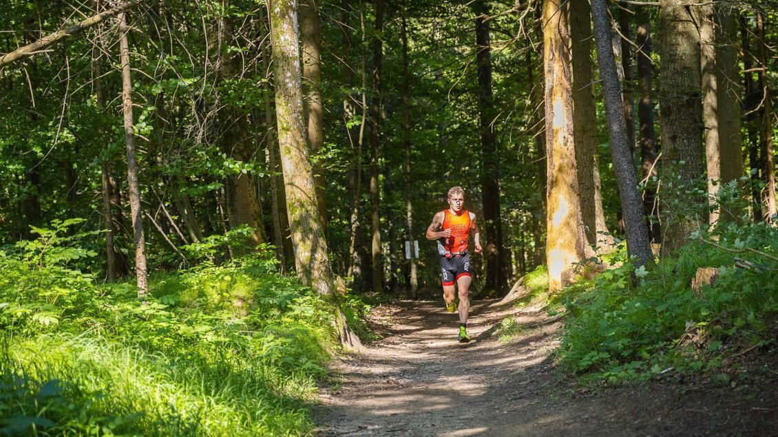 <p>Ein sportlicher Läufer vom Xterra Austria Triathlon läuft auf einem schmalen Weg durch einen grünen Wald in Altaussee. Die Sonne scheint durch die Bäume und beleuchtet die Landschaft.</p> | © XTERRA Austria