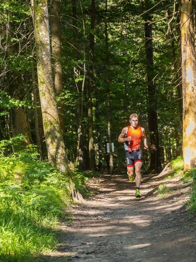 <p>Ein sportlicher Läufer vom Xterra Austria Triathlon läuft auf einem schmalen Weg durch einen grünen Wald in Altaussee. Die Sonne scheint durch die Bäume und beleuchtet die Landschaft.</p> | © XTERRA Austria
