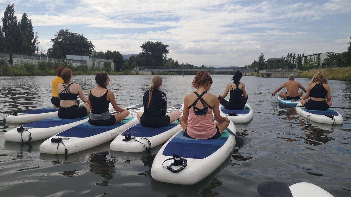Eine Gruppe von Menschen paddelt auf Stand-Up-Paddle-Boards auf einem ruhigen Wasser. Die Umgebung ist grün mit Bäumen und einem klaren Himmel.