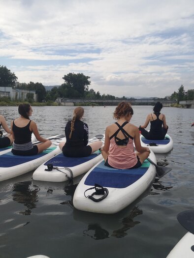 Eine Gruppe von Menschen paddelt auf Stand-Up-Paddle-Boards auf einem ruhigen Wasser. Die Umgebung ist grün mit Bäumen und einem klaren Himmel.