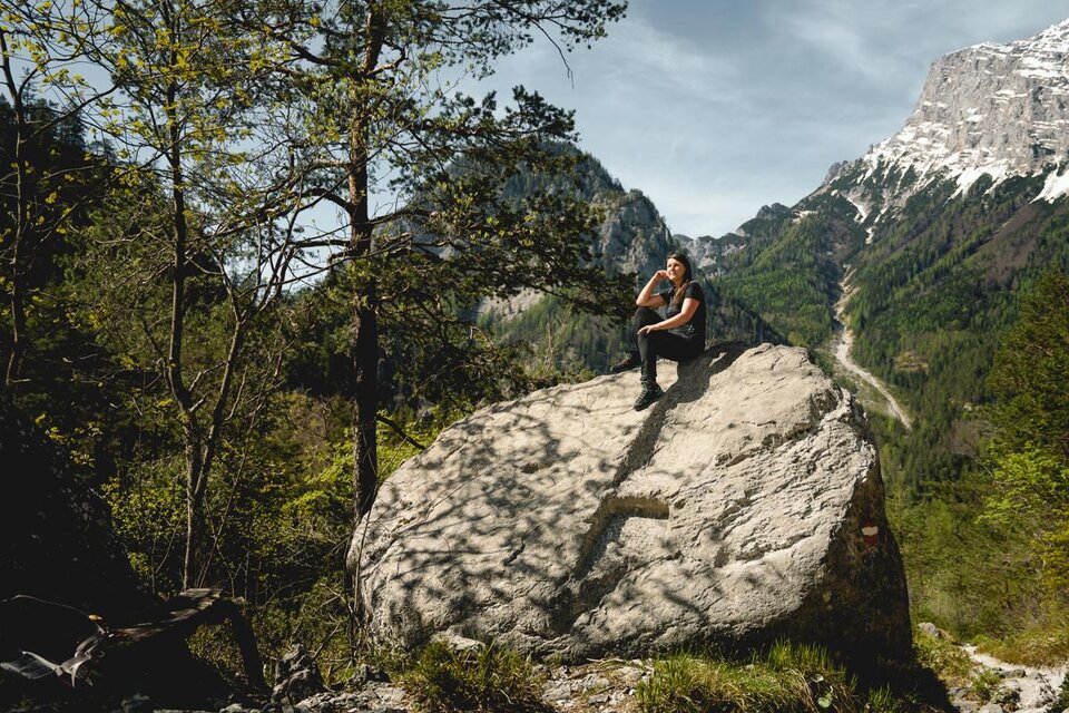 Ein Wanderer sitzt auf einem großen Felsen in einer malerischen Berglandschaft. Die Umgebung ist grün, mit Bäumen und hohen Bergen im Hintergrund. | © Tourismusverband Gesäuse