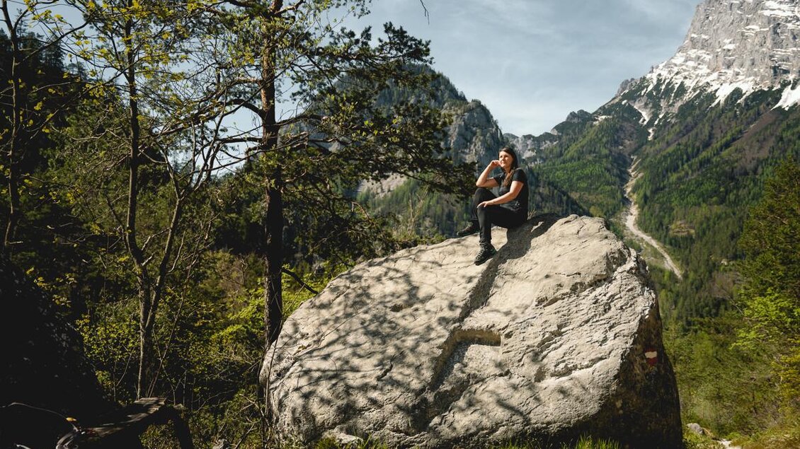 Ein Wanderer sitzt auf einem großen Felsen in einer malerischen Berglandschaft. Die Umgebung ist grün, mit Bäumen und hohen Bergen im Hintergrund. | © Tourismusverband Gesäuse