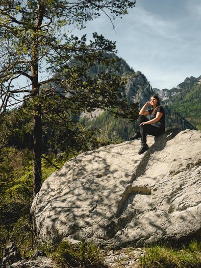 Ein Wanderer sitzt auf einem großen Felsen in einer malerischen Berglandschaft. Die Umgebung ist grün, mit Bäumen und hohen Bergen im Hintergrund. | © Tourismusverband Gesäuse