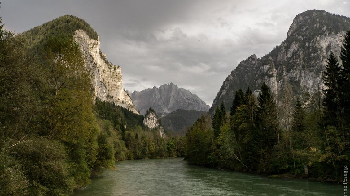 Eine ruhige Landschaft mit einem fließenden Fluss, umgeben von hohen Bergen und dichten Wäldern. Der Himmel ist bewölkt und verleiht der Szene eine dramatische Stimmung.