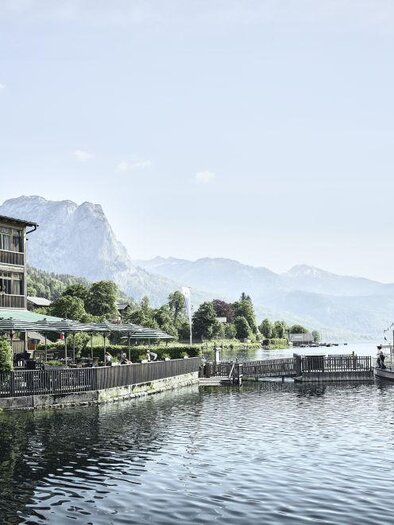 Ein malerisches Seeufer mit einem charmanten Boot und einer Gaststätte. Im Hintergrund erheben sich majestätische Berge unter einem klaren Himmel. | © www.tauroa.at