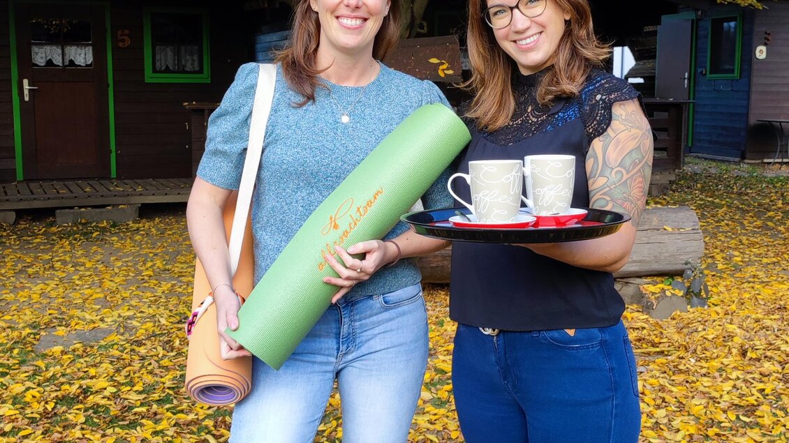 Zwei Frauen stehen in einem herbstlichen Park voller bunter Blätter. Eine hält eine Yogamatte und die andere ein Tablett mit Tassen. | © Marlies Eichelberger