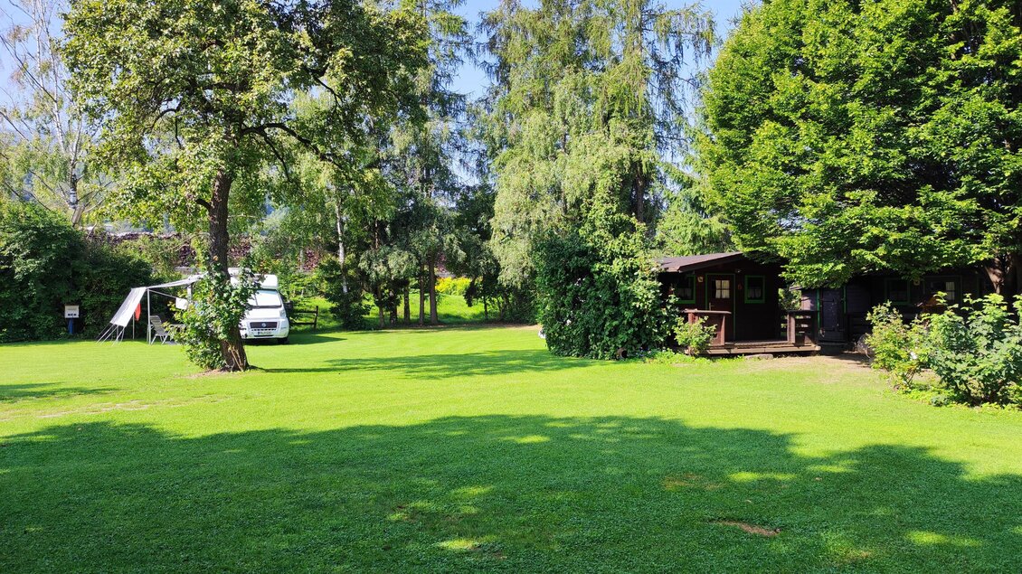 Eine grüne Wiese mit Bäumen und einem kleinen Holzhaus. Im Hintergrund ist ein Wohnmobil zu sehen. | © Landhaus Camping Pichler