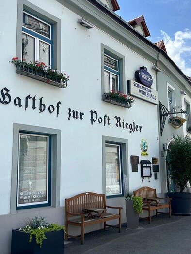 Ein älteres Gasthaus mit dem Namen "Gasthof zur Post Riegler" in einer malerischen Straße. Die Fassade ist hell gestrichen und mit Blumen in Fensterkästen geschmückt. | © TV Hochsteiermark
