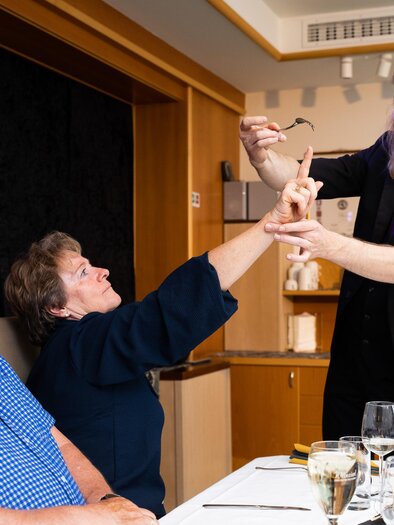 A magician entertains an audience at the table. A woman reaches for a trick while a man watches.