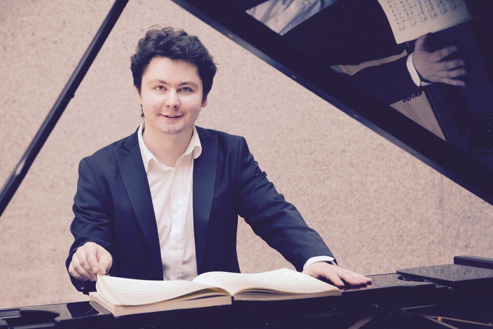 Ein junger Pianist sitzt am Klavier und blickt lächelnd in die Kamera. Vor ihm liegt ein aufgeschlagenes Notenbuch. | © A. Bashynsky
