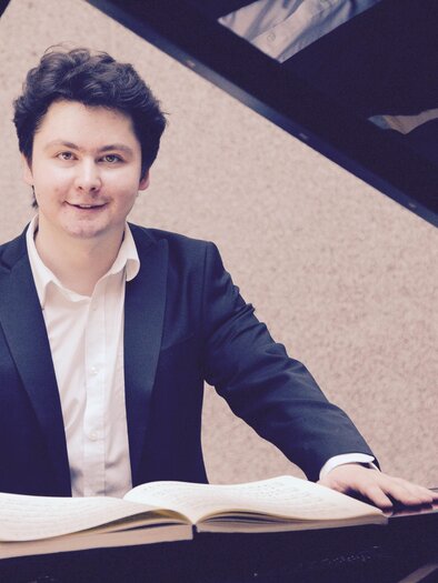 Ein junger Pianist sitzt am Klavier und blickt lächelnd in die Kamera. Vor ihm liegt ein aufgeschlagenes Notenbuch. | © A. Bashynsky