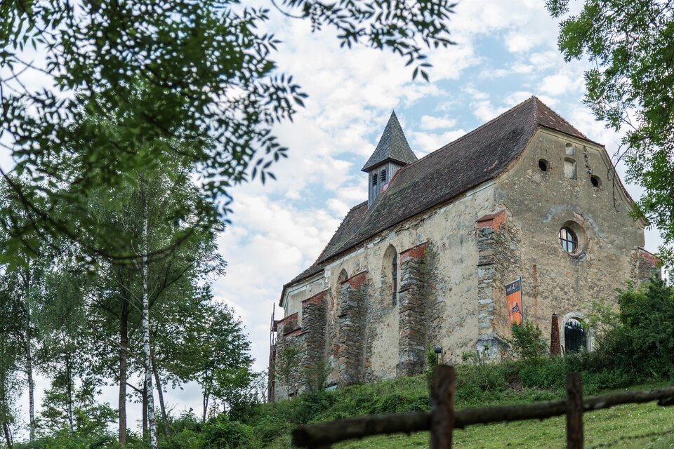 Eine alte Kirche umgeben von Bäumen und Wiesen. Der Himmel ist teilweise bewölkt und die Architektur ist rustikal. | © fotodesign.at by Marcus Auer