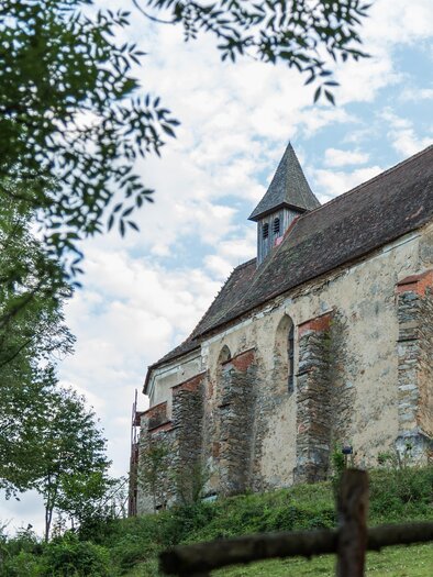 Eine alte Kirche umgeben von Bäumen und Wiesen. Der Himmel ist teilweise bewölkt und die Architektur ist rustikal. | © fotodesign.at by Marcus Auer