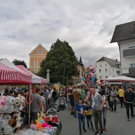 Ein belebter Markt mit vielen Menschen und bunten Ständen. Im Hintergrund sind Häuser und ein Himmel mit grauen Wolken zu sehen. | © Stadtgemeinde Fürstenfeld