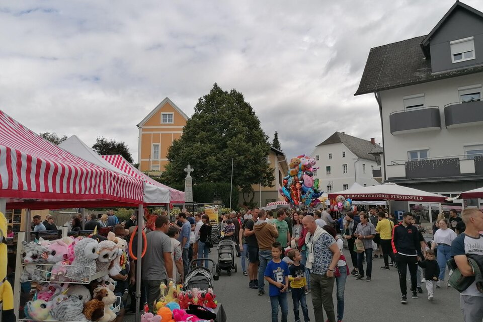 Ein belebter Markt mit vielen Menschen und bunten Ständen. Im Hintergrund sind Häuser und ein Himmel mit grauen Wolken zu sehen. | © Stadtgemeinde Fürstenfeld