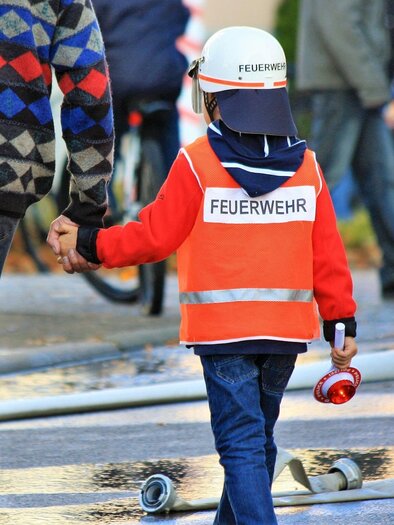 Ein Kind in einer Feuerwehrjacke hält die Hand eines Erwachsenen. Das Kind trägt einen Feuerwehrhelm und hat eine kleine Sirene in der anderen Hand.