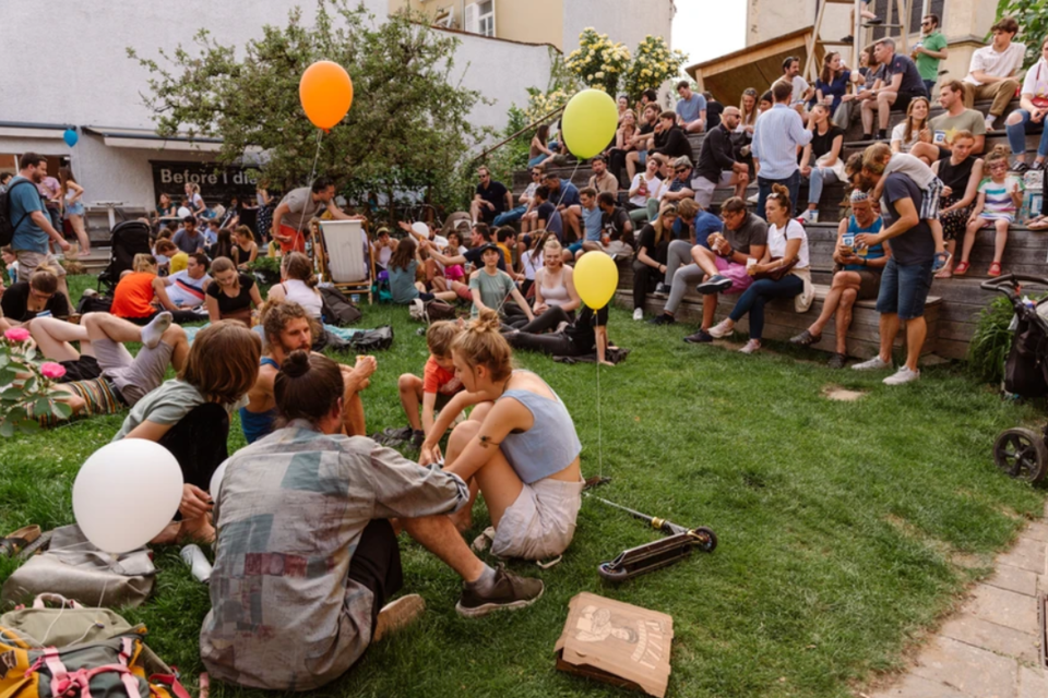 Eine lebhafte Veranstaltung im Freien mit vielen Menschen, die auf dem Gras sitzen und sich unterhalten. Einige halten Luftballons, während andere auf einer Bühne oder Treppe stehen. | © _LENA BALOCH_