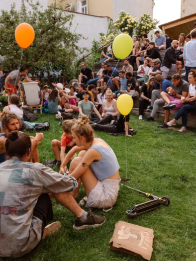 A lively outdoor event with many people sitting on the grass and chatting. Some are holding balloons, while others are standing on a stage or staircase. | © _LENA BALOCH_